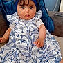 baby, child, dress, headband, couch, cushion, indoor, portrait, person, blue, white, pattern, leaf, plant, sitting, curly_hair, face, eyes, hand, cute