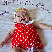 Olivia is registered to the contest to win money with this photo: baby, infant, smiling, red_dress, polka_dots, headband, flower, blanket, lying_down, white_blanket, happy, cute, child, person, footwear, sock, indoors, celebration, joy, adorable