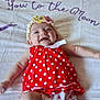 baby, infant, smiling, red_dress, polka_dots, headband, flower, blanket, lying_down, white_blanket, happy, cute, child, person, footwear, sock, indoors, celebration, joy, adorable