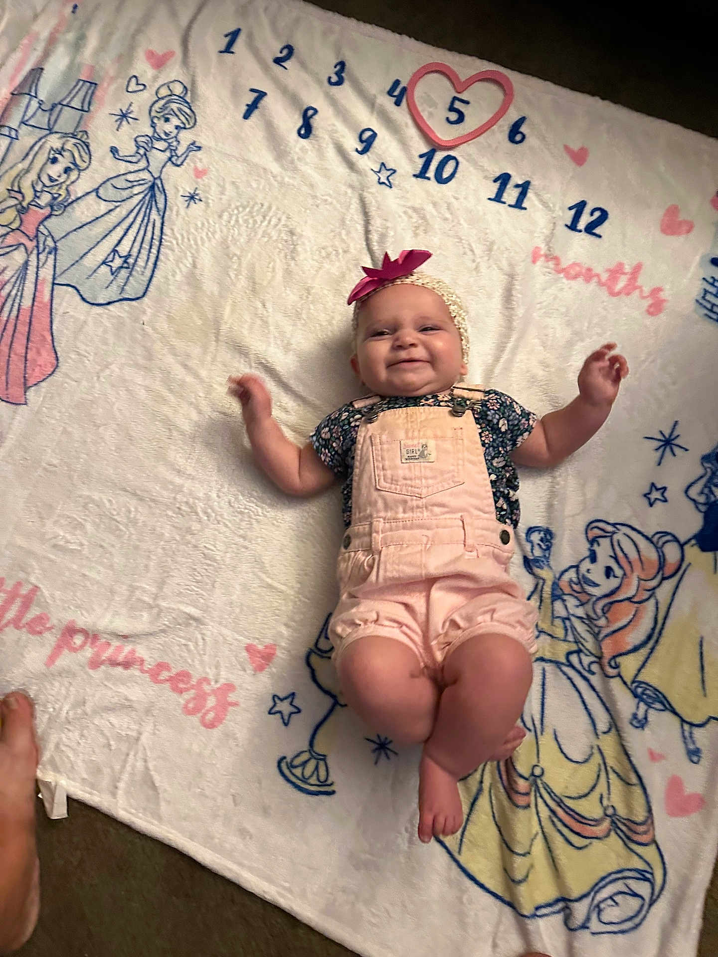 Jacqueline is registered to the contest to win money with this photo: baby, girl, smiling, blanket, princess, overalls, headband, bow, numbers, months, cartoon, character, lying_down, happy, cute, indoor, carpet, feet, child, portrait