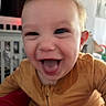 baby, blurred_background, child, closeup, crib, cute, expression, face, happy, head, home, indoor, infant, joy, mouth_open, mustard_outfit, person, smiling, tongue_out, young_child