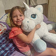 Delilah joined the competition — help win amazing prizes! bed, bedroom, blanket, brown_hair, child, family_photo, girl, happy, hugging, indoors, pillow, pink_shirt, playtime, plush_toy, portrait, shorts, smiling, stuffed_animal, toy, unicorn