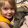 animal, bed, bedding, blanket, brown_hair, child, closeup, companion, cuddle, dog, family, girl, happy, hugging, indoor, pet, pillow, portrait, smiling, yellow_shirt