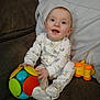 baby, child, smiling, toy, ball, couch, onesie, sitting, happy, indoor, face, infant, play, cute, person, footwear, colorful, soft_toy, blanket, home