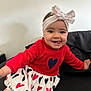 baby, toddler, headband, bow, smile, tongue_out, red_sweater, heart_skirt, skirt, sitting, sofa, black_sofa, indoor, white_wall, happy, face, eyes, hand, portrait, clothing