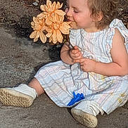 Lyra is registered to the contest to win money with this photo: toddler, child, flower, outdoor, garden, smile, dress, shoes, plant, nature, sitting, cute, happy, person, young_child, blossom, sunlight, daytime, curly_hair, holding
