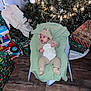 baby, infant, christmas_tree, christmas_lights, presents, gift_wrap, holiday, onesie, baby_chair, flooring, wood_floor, decorations, pine_cones, ornaments, green_blanket, socks, indoors, festive, child, cozy
