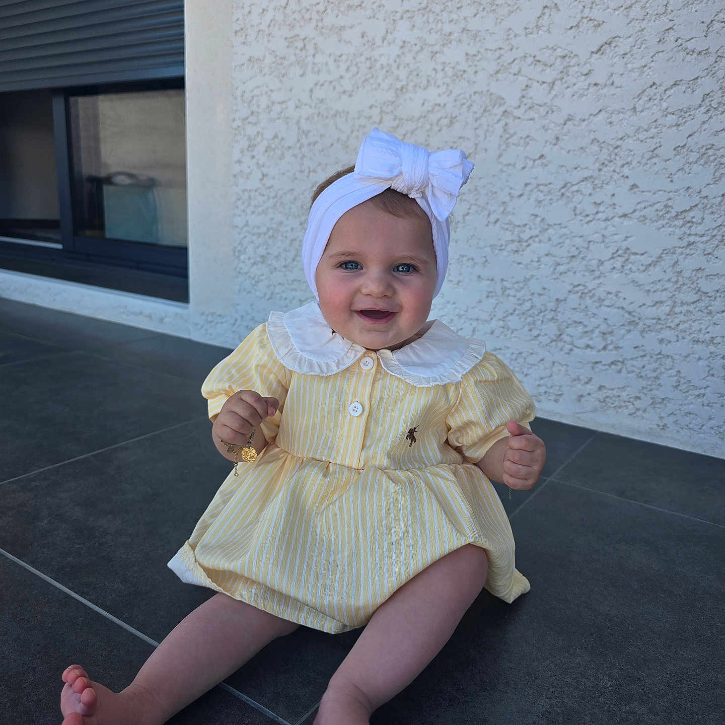 Rachel a rejoint le concours — aidez-le/la à gagner de superbes lots ! baby, child, smiling, yellow_dress, headband, bow, blue_eyes, sitting, floor, tile, outdoor, wall, window, happy, cute, person, portrait, clothing, infant, baby_girl