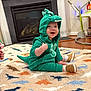 baby, child, dinosaur_costume, green, onesie, smiling, sitting, rug, pattern, dinosaur, indoor, floor, shoe, toy, fireplace, home, happy, cute, infant, playful