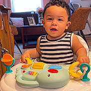 Owen is registered to the contest to win money with this photo: toddler, child, baby_walker, toy, striped_clothing, indoor, living_room, wooden_floor, curly_hair, smiling, furniture, chair, table, curtains, window, foot, person, playtime, happy, home