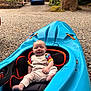 baby, kayak, blue_kayak, gravel, driveway, stone_house, car, blue_car, tree_trunk, greenery, outdoor, smiling, child, seat, playful, nature, daylight, candid, happy, baby_clothes