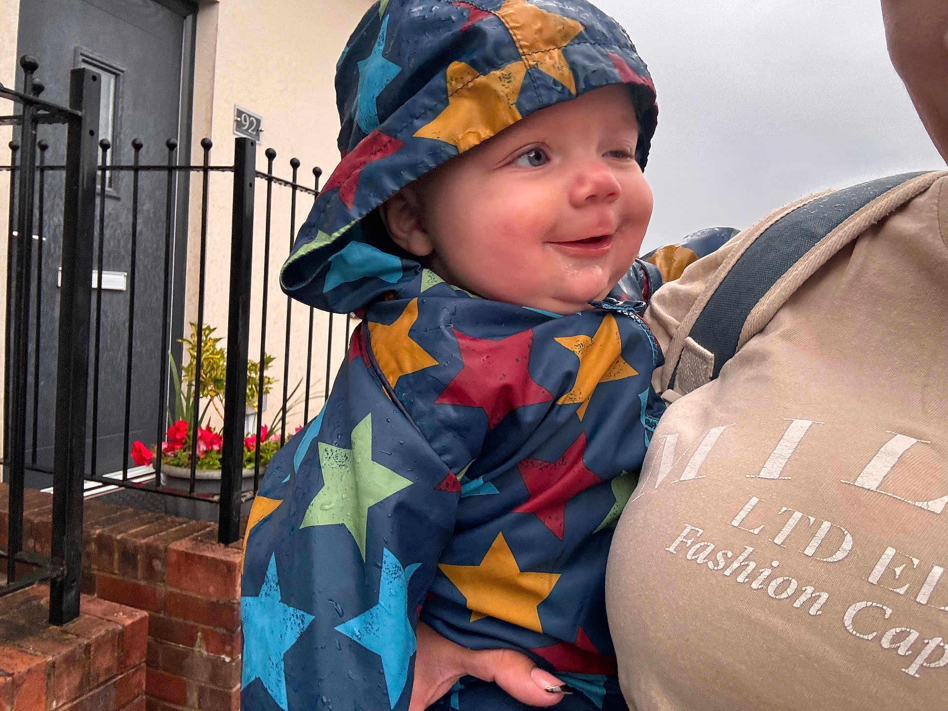 Otto is registered to the contest to win money with this photo: baby, child, raincoat, stars, hood, smile, winking, raindrops, adult, holding, hand, door, fence, flowers, plants, brick, outdoor, clothing, portrait, happy