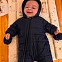 Otto is registered to the contest to win money with this photo: baby, infant, snowsuit, black_clothing, hood, smile, lying_down, changing_pad, animal_print, wooden_table, indoor, cute, child, happy, mittens, face, person, portrait, baby_clothes, cozy