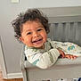 toddler, child, crib, smiling, curly_hair, onesie, dinosaur_print, baby, indoor, wooden_floor, bedding, animal_print, happy, face, person, furniture, home, cute, playful, standing