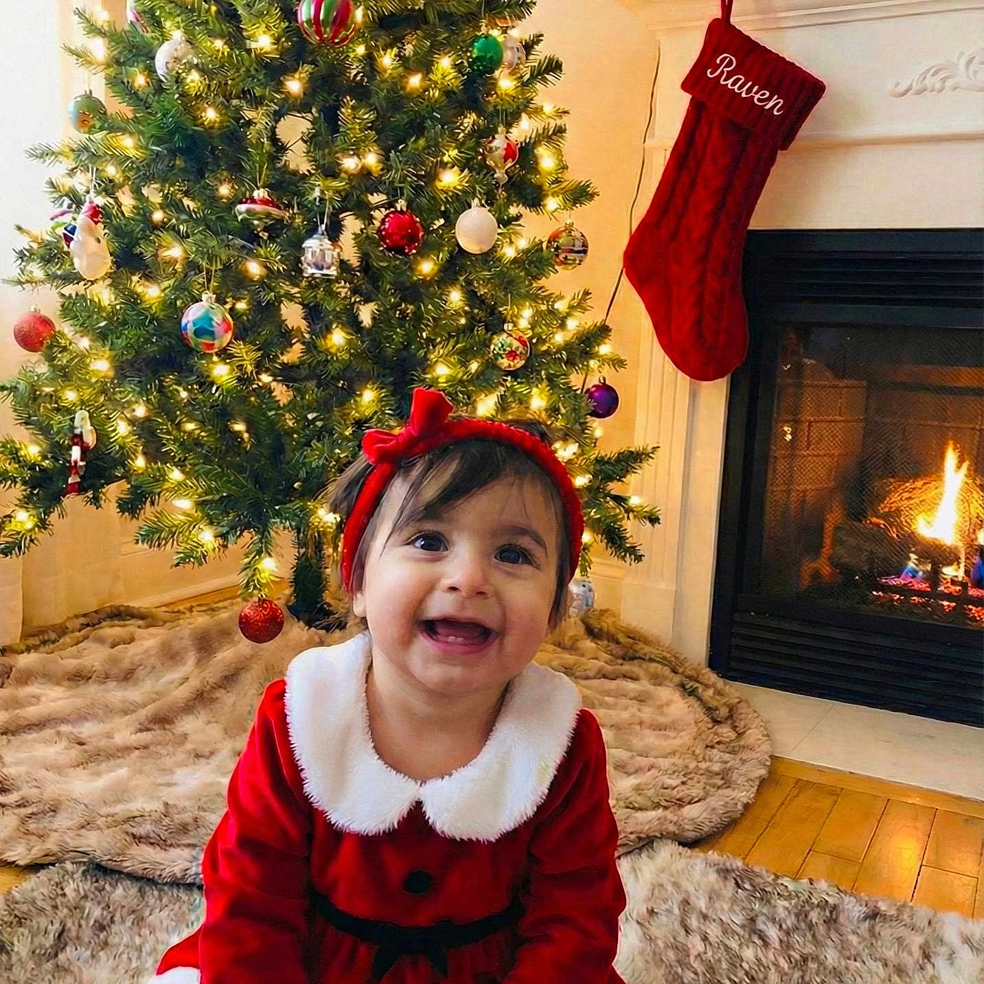 Raven is registered to the contest to win money with this photo: carpet, celebration, child, christmas_tree, cozy, decorations, festive_clothing, fireplace, happy, headband, holiday, home, indoors, lights, red_dress, seasonal, smiling, stocking, toddler, warmth