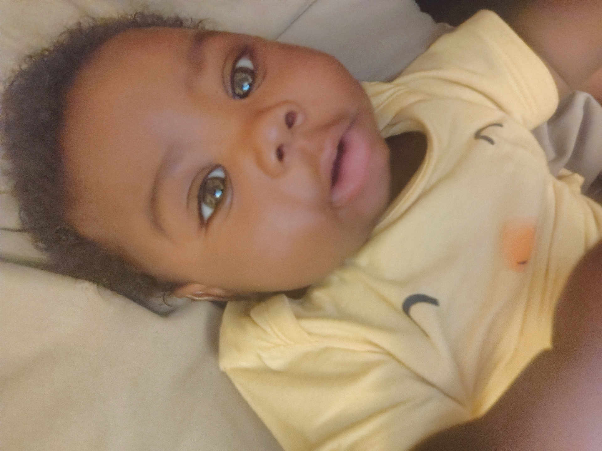 Jamarious joined the competition — help win amazing prizes! baby, child, face, eyes, mouth, yellow_shirt, skin, hair, lying_down, portrait, infant, person, closeup, expression, cute, young_child, indoors, soft_lighting, casual_clothing, human