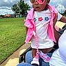 child, toddler, pink_sunglasses, pink_vest, white_tshirt, necklace, sneakers, grass, outdoor, cloudy_sky, adult_hand, jeans, stroller, sidewalk, fashion, cute, person, accessories, summer, casual