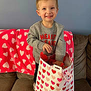 Karson joined the competition — help win amazing prizes! child, toddler, boy, smiling, heart_pattern, gift_bag, couch, blanket, gray_sweater, living_room, portrait, standing, present, valentine, hearts, pink, red, joyful, playful, indoor