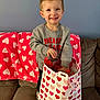 child, toddler, boy, smiling, heart_pattern, gift_bag, couch, blanket, gray_sweater, living_room, portrait, standing, present, valentine, hearts, pink, red, joyful, playful, indoor