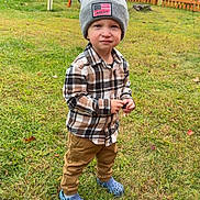 Karson is registered to the contest to win money with this photo: child, toddler, beanie, hat, plaid_shirt, flannel, grass, yard, fence, autumn, leaves, crocs, shoes, portrait, standing, smirk, face, person, outdoor, casual_clothing