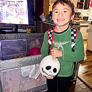 Liam joined the competition — help win amazing prizes! backpack, boy, casual_clothing, child, curly_hair, decorated_pumpkin, furniture, green_shirt, happy, holding, home, indoor, kitchen, person, playpen, pumpkin, smile, television, toy, wood_floor