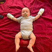 Tobias is registered to the contest to win money with this photo: baby, sleeping, pacifier, mittens, onesie, bed, red_bedspread, speaker, jbl, remote_control, blanket, infant, indoors, resting, cute, child, comfort, soft_lighting, relaxed, portrait