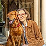 dog, golden_retriever, woman, glasses, brown_jacket, jeans, smile, sunlight, outdoor, architecture, stone_columns, archway, friendship, pet, happy, portrait, sitting, tongue_out, red_collar, casual_clothing