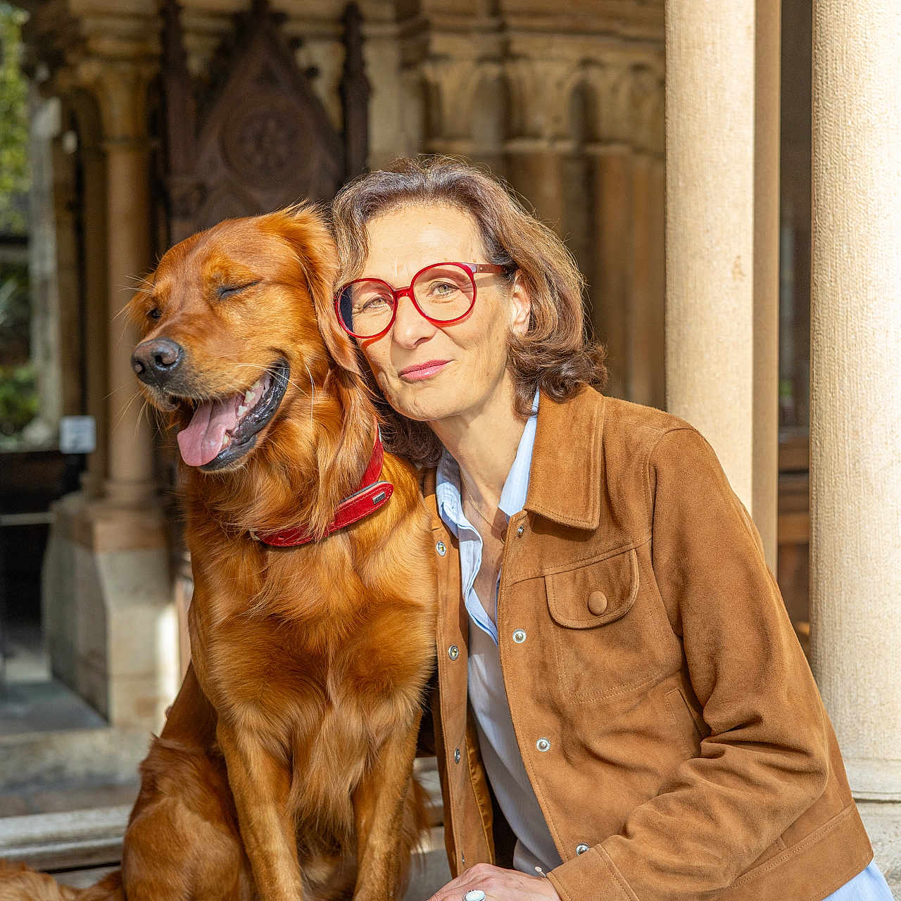 Ugo participe au concours pour gagner de l'argent avec cette photo : dog, golden_retriever, woman, glasses, brown_jacket, jeans, smile, sunlight, outdoor, architecture, stone_columns, archway, friendship, pet, happy, portrait, sitting, tongue_out, red_collar, casual_clothing