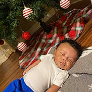 Keegan is registered to the contest to win money with this photo: baby, sleeping, blanket, christmas_tree, ornaments, red, white, green, plaid, floor, wooden_floor, clothing, pants, shirt, holiday, festive, indoor, decorations, peaceful, person