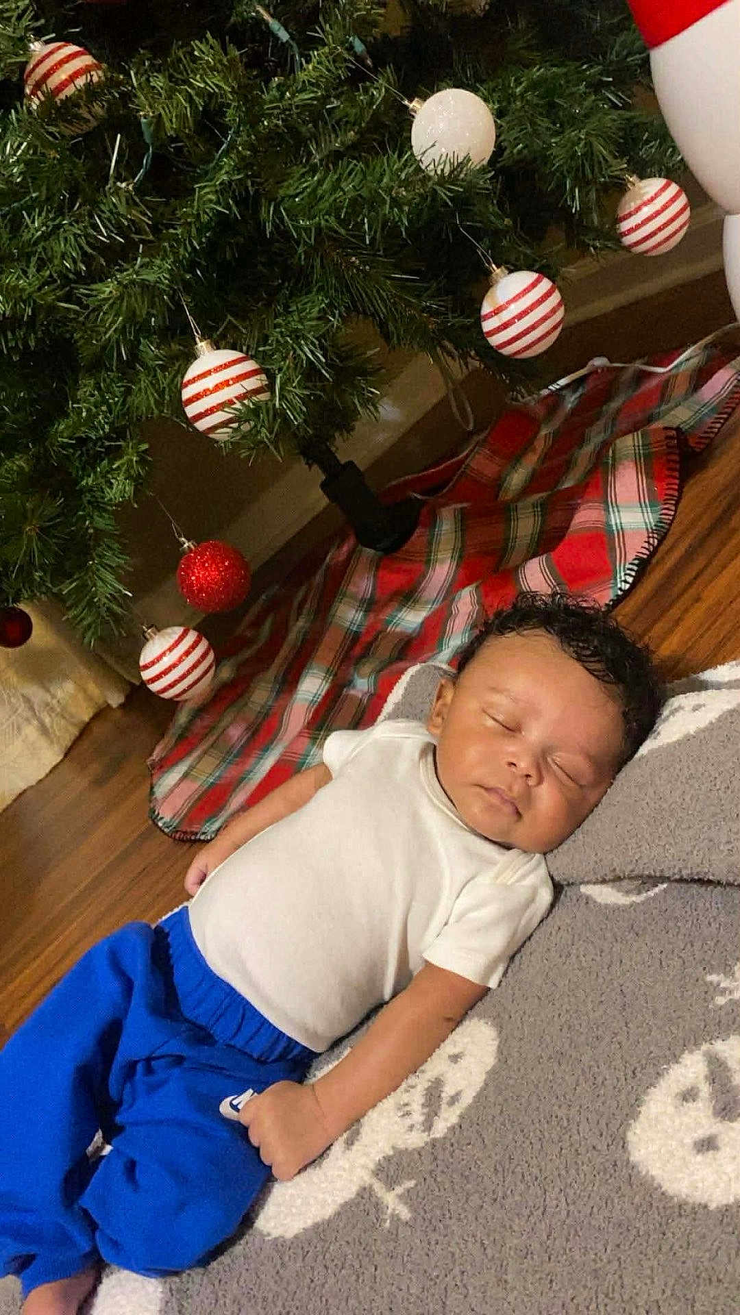 Keegan is registered to the contest to win money with this photo: baby, sleeping, blanket, christmas_tree, ornaments, red, white, green, plaid, floor, wooden_floor, clothing, pants, shirt, holiday, festive, indoor, decorations, peaceful, person