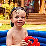 backward_hat, bare_chest, boy, cap, child, daylight, fish_toy, garden, happy, inflatable_pool, outdoor, playing, pool, red_toy, skin, smiling, summer, toddler, toy, water