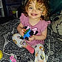 bedspread, blanket, blue_eyes, child, colorful_toy, curly_blonde_hair, curly_hair, cute, happy, indoor, pacifier, pajamas, pink_shirt, play, room, sitting, smiling, toddler, toy, young_child