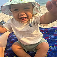 Noah is registered to the contest to win money with this photo: baby, child, smiling, hat, bucket_hat, beach, ocean, sand, blanket, sitting, happy, cute, infant, sunlight, outdoor, person, clothing, foot, hand, portrait