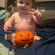 Carter is registered to the contest to win money with this photo: baby, child, pumpkin, jack_o_lantern, bed, blanket, toddler, carved_pumpkin, indoor, portrait, cute, smile, hair, face, halloween, decor, tapestry, furniture, room, expression