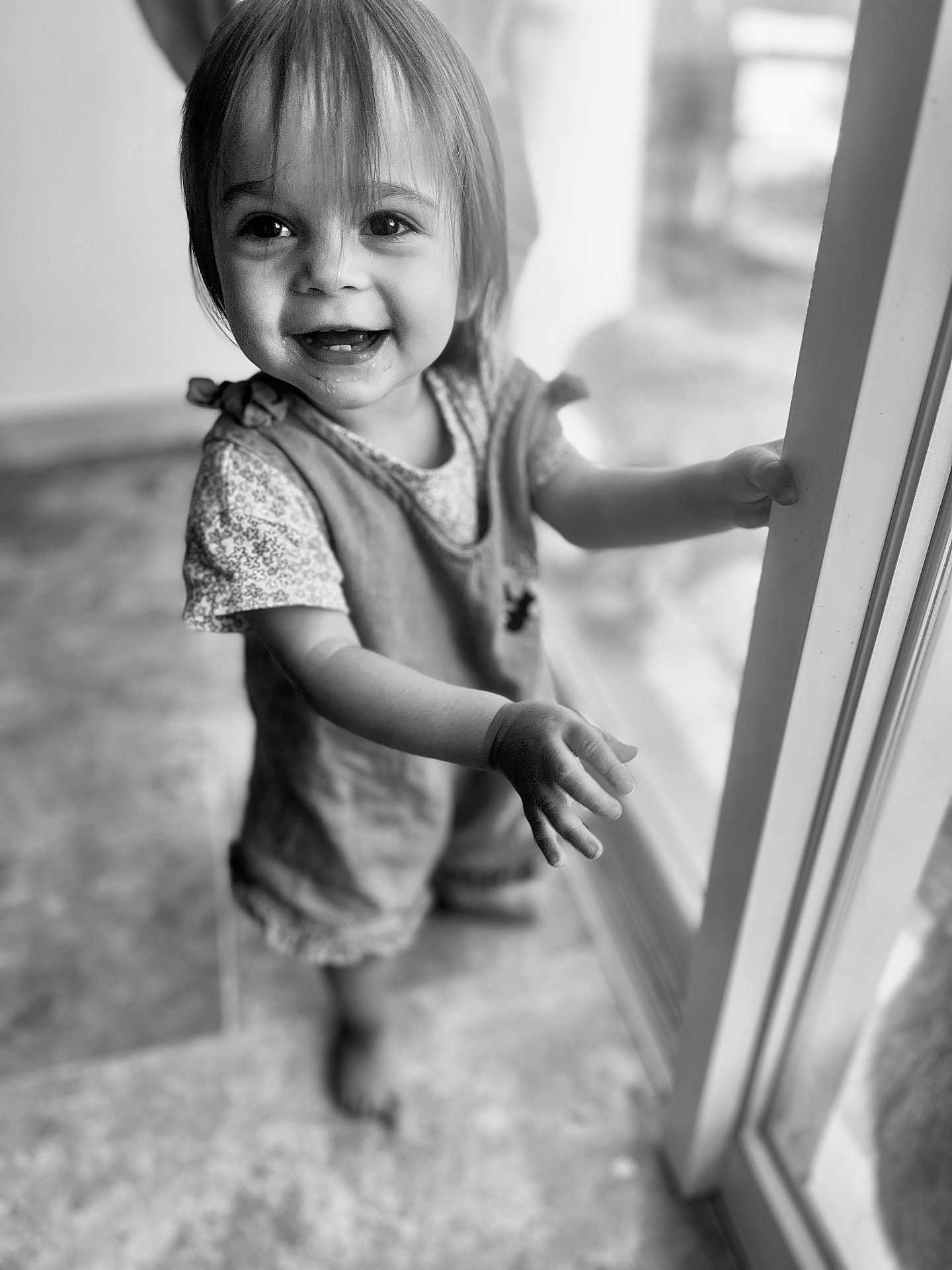 Hanaé participe au concours pour gagner de l'argent avec cette photo : toddler, child, smiling, indoor, black_and_white, portrait, happy, face, hand, glass_door, clothing, floor, person, cute, young_child, expression, window, hair, barefoot, light