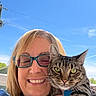 woman, cat, tabby_cat, glasses, smile, outdoor, blue_sky, sun_decoration, happy, pet, face, portrait, holding, feline, animal, person, daylight, nature, closeup, casual