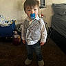 bed, blanket, carpet, casual, child, clothing, container, curious, floor, furniture, indoor, pacifier, person, plastic_bottle, standing, striped_shirt, toddler, toy_storage, vest, young_child