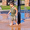 Sabastian joined the competition — help win amazing prizes! child, water, splash_pad, barefoot, summer, outdoor, play, wet_clothes, tank_top, pants, brick_pillar, black_pole, grass, car, reflection, sunlight, water_spray, happy, fun, daytime