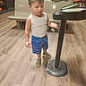 activity, background_people, bench, bowling_alley, bowling_balls, casual_clothing, child, curious, floor, gaming_machine, indoor, leisure, people, shorts, sneakers, standing, tank_top, toddler, wood_floor, young_child