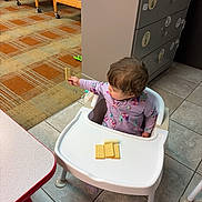 Raven Wright is registered to the contest to win money with this photo: toddler, child, high_chair, cracker, purple_onesie, floor_tiles, cabinet, crib, carpet, table, room, indoor, baby, furniture, childcare, snack, curious, seated, holding, playroom