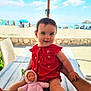 baby, child, doll, table, hand, beach, umbrella, ocean, sky, clouds, sunny, red_dress, toy, person, outdoor, summer, sand, chair, smile, portrait