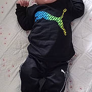 Thomas is registered to the contest to win money with this photo: baby, infant, child, black_clothing, puma_logo, bedspread, quilt, white_background, small_red_flowers, lying_down, looking_up, curious_expression, long_sleeves, pants, footwear_absent, indoor, soft_lighting, portrait, person, cute