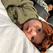 Lillyanna joined the competition — help win amazing prizes! baby, child, headband, olive_green_clothing, white_blanket, face, lying_down, indoor, fan, laundry_basket, curious_expression, person, sibling, home, soft_lighting, closeup, portrait, young_child, family, cozy