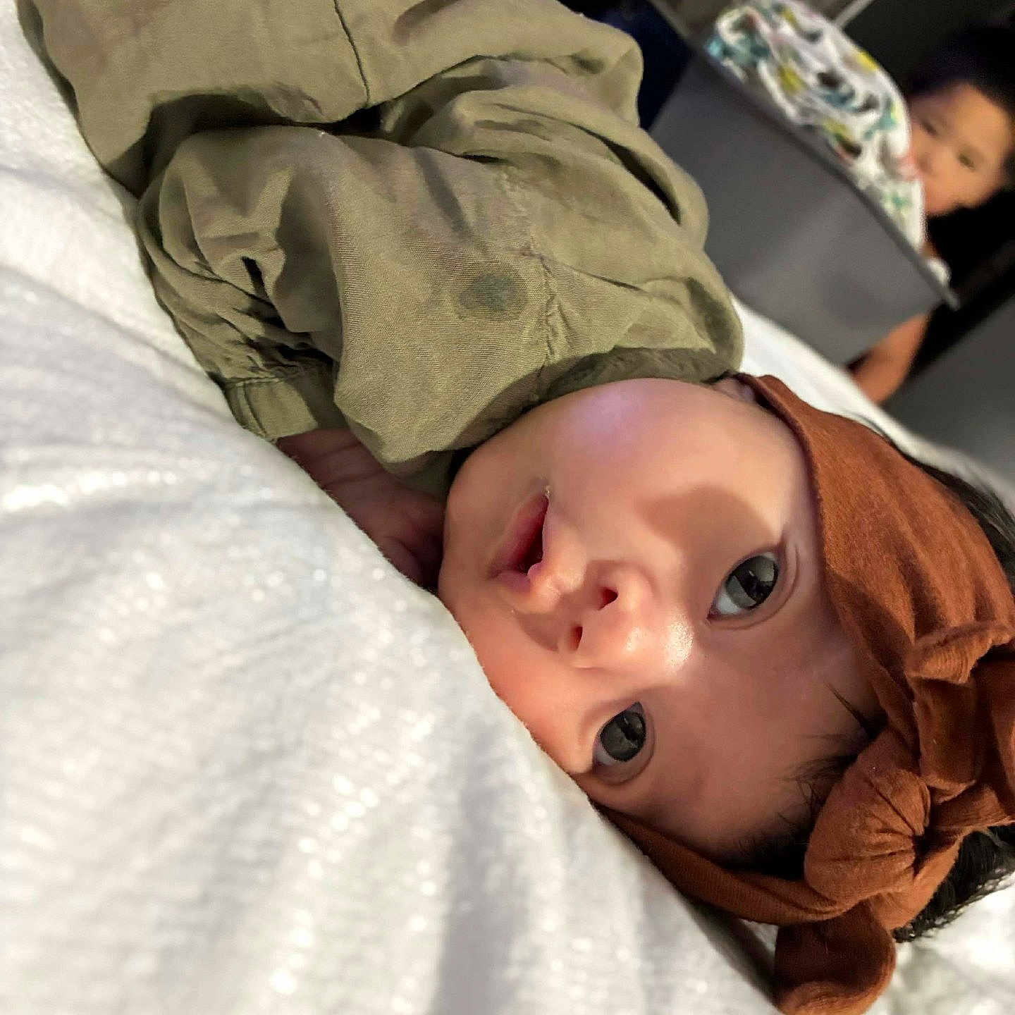 Lillyanna joined the competition — help win amazing prizes! baby, child, headband, olive_green_clothing, white_blanket, face, lying_down, indoor, fan, laundry_basket, curious_expression, person, sibling, home, soft_lighting, closeup, portrait, young_child, family, cozy