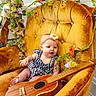 baby, child, ukulele, armchair, velvet, yellow, flower, headband, blue_clothing, checkered, wood, musical_instrument, indoor, cute, sitting, decor, greenery, playful, infant, portrait
