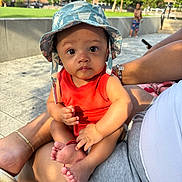 Noah joined the competition — help win amazing prizes! baby, child, hat, orange_clothing, outdoor, person, sitting, legs, hand, foot, adult, bracelet, pavement, background, greenery, building, sunlight, summer, casual, playful