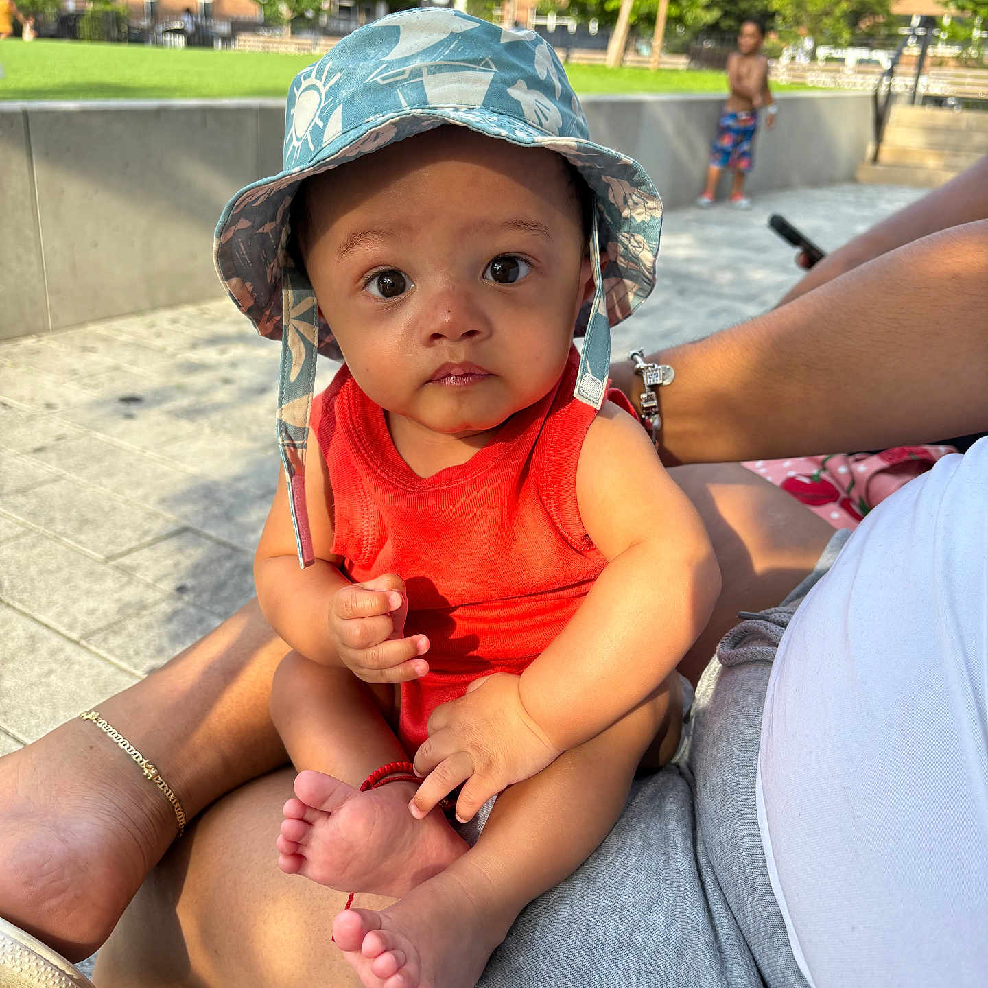 Noah joined the competition — help win amazing prizes! adult, baby, background, bracelet, building, casual, child, foot, greenery, hand, hat, legs, orange_clothing, outdoor, pavement, person, playful, sitting, summer, sunlight