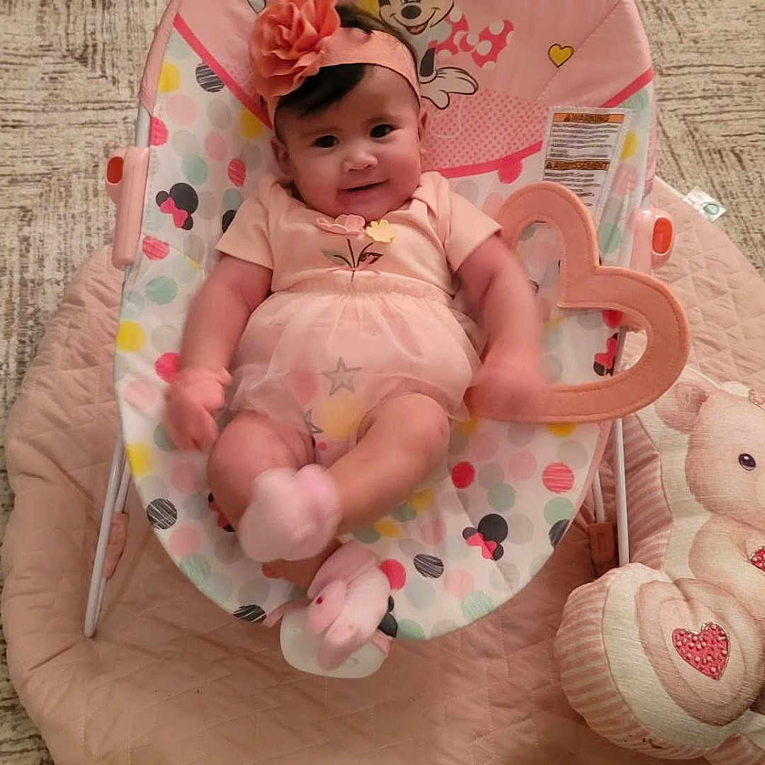 Kindra is registered to the contest to win money with this photo: baby, baby_rocker, carpet, child, colorful, cute, hand, headband, heart_toy, indoor, infant, minnie_mouse, person, pink_clothing, playful, quilt, seated, smartphone, smiling, soft_toy