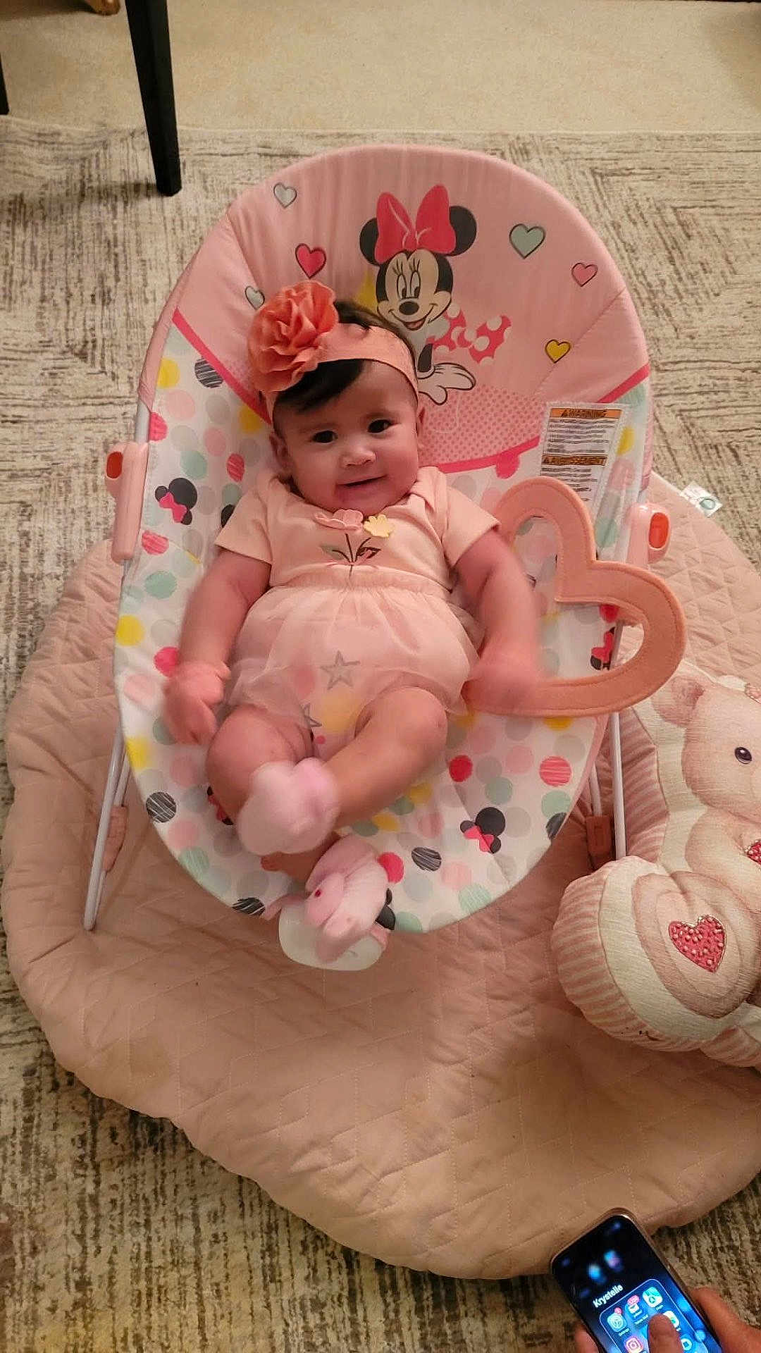 Kindra is registered to the contest to win money with this photo: baby, baby_rocker, carpet, child, colorful, cute, hand, headband, heart_toy, indoor, infant, minnie_mouse, person, pink_clothing, playful, quilt, seated, smartphone, smiling, soft_toy