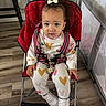 toddler, child, stroller, red, bow, curly_hair, heart_pattern, white_shoes, indoors, wooden_floor, baby_crib, clothing, person, face, seat_belt, expression, cute, small_hand, sitting, curious